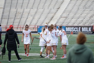 A group of female lacrosse players is standing together on a sports field, seemingly strategizing before or during a game. They are wearing team uniforms in white with red accents, and some are holding lacrosse sticks. In the background, there is a spectator wearing a jacket and cap, as well as rows of empty stadium seats. There is also a large advertisement displayed.