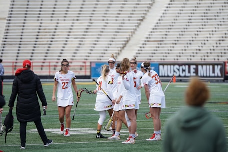 A group of female lacrosse players is standing together on a sports field, seemingly strategizing before or during a game. They are wearing team uniforms in white with red accents, and some are holding lacrosse sticks. In the background, there is a spectator wearing a jacket and cap, as well as rows of empty stadium seats. There is also a large advertisement displayed.