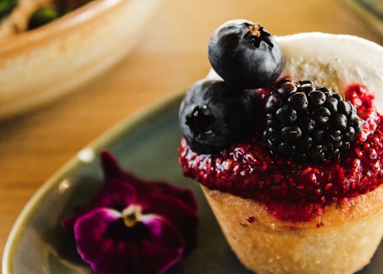 A close-up of a dessert featuring a muffin topped with vibrant red berry compote and fresh berries, including blueberries and a blackberry. A purple flower is placed on the plate beside the muffin.
