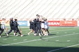 A group of athletes are celebrating on a lacrosse field, with some wearing black jackets and others in white uniforms. The bleachers in the background are mostly empty, and the turf is marked for sports activities.