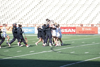 A group of athletes are celebrating on a lacrosse field, with some wearing black jackets and others in white uniforms. The bleachers in the background are mostly empty, and the turf is marked for sports activities.