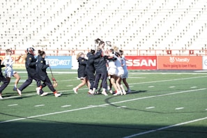 A group of athletes are celebrating on a lacrosse field, with some wearing black jackets and others in white uniforms. The bleachers in the background are mostly empty, and the turf is marked for sports activities.