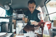 A man is preparing coffee using a pour-over method in a cozy, compact space, likely a van or small shop. The interior is organized with various coffee-making equipment, including a drip coffee set, a grinder, and multiple containers. The atmosphere is intimate and focused on the craft of coffee preparation.