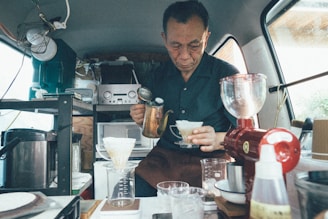A man is preparing coffee using a pour-over method in a cozy, compact space, likely a van or small shop. The interior is organized with various coffee-making equipment, including a drip coffee set, a grinder, and multiple containers. The atmosphere is intimate and focused on the craft of coffee preparation.