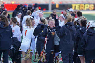 A group of female lacrosse players, some in white sports uniforms and others in black jackets, gather on the field. They appear to be celebrating and engaging in conversation, with one player prominently smiling and holding a lacrosse stick. The background includes a sports stadium with red seating and a blurry banner featuring the word 'TERPS'.