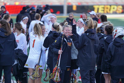 A group of female lacrosse players, some in white sports uniforms and others in black jackets, gather on the field. They appear to be celebrating and engaging in conversation, with one player prominently smiling and holding a lacrosse stick. The background includes a sports stadium with red seating and a blurry banner featuring the word 'TERPS'.