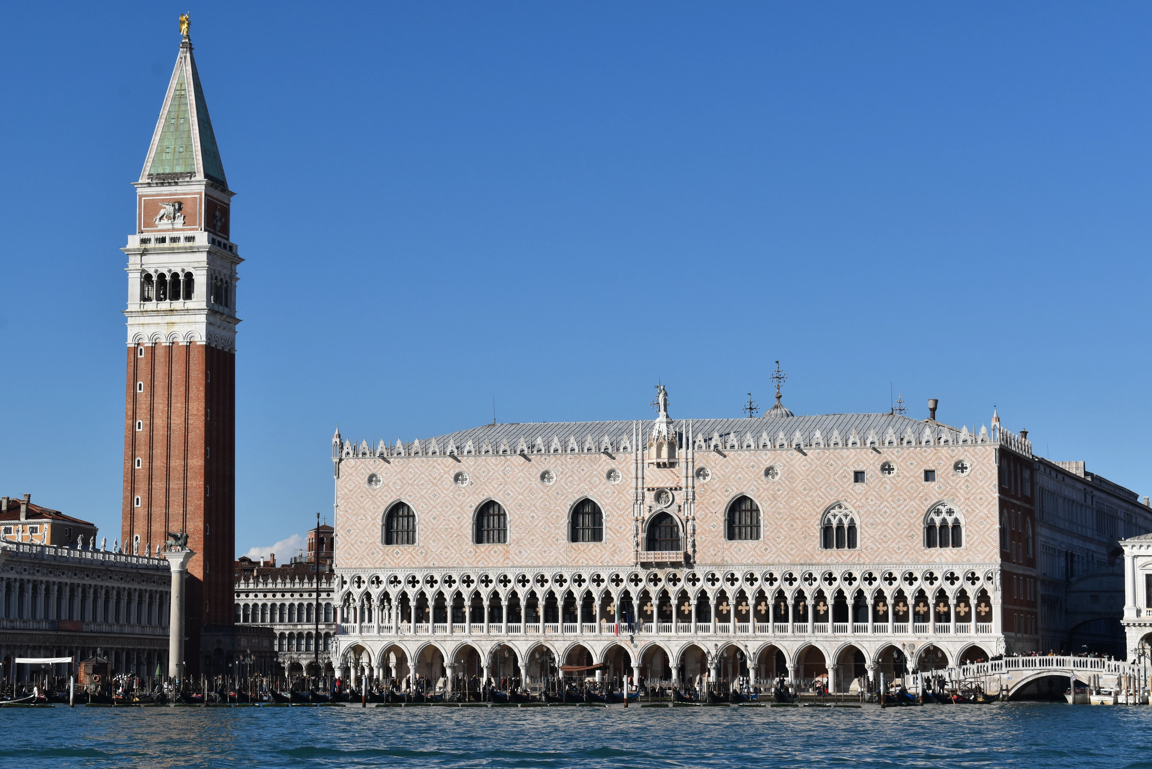 The Doge's Palace and the brick campanile of St Mark's seen across the water in Venice under a cloudless blue sky, gondolas moored along the waterfront arcade