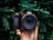 A hand is holding a Nikon camera surrounded by lush green foliage. The camera has a clear, wide lens, and the scene provides a striking contrast between the mechanical equipment and the organic environment.