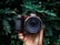 A hand is holding a Nikon camera surrounded by lush green foliage. The camera has a clear, wide lens, and the scene provides a striking contrast between the mechanical equipment and the organic environment.