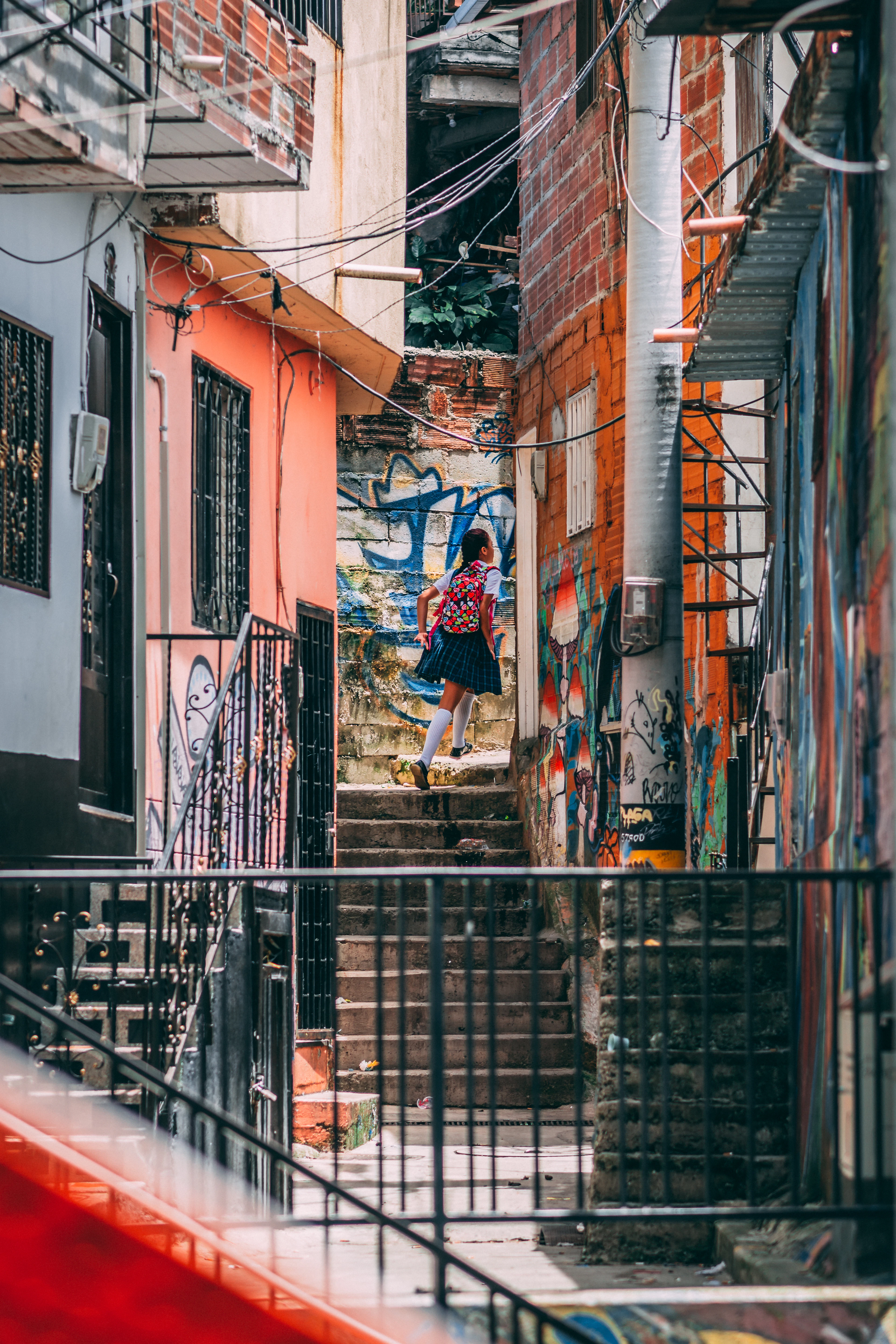A schoolgirl in uniform with a flowered backpack climbing a steep concrete staircase in Medellin's Comuna 13, between brick and pink-painted houses whose walls are covered in blue and orange spray-painted murals.