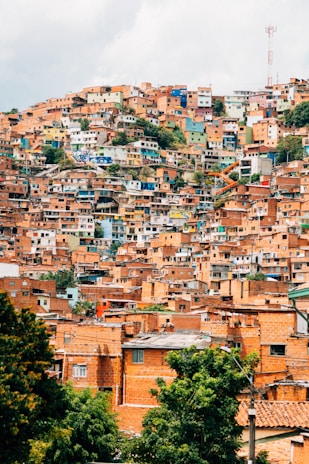 A densely packed hillside neighborhood with numerous multicolored houses built closely together. The vibrant facades include shades of orange, blue, green, and beige. Some greenery is visible among the buildings, and a communication tower is perched at the top of the hill.