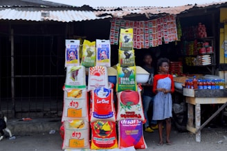 A small market stall displays various bags of rice, stacked in multiple columns. Two people, a young boy and a girl, are present; the boy holds a bowl of grains. Several red bottles are neatly arranged on a wooden table. Packs of hanging seasonings and instant noodles are visible in the background inside the dimly lit stall, with a metal roof overhead.
