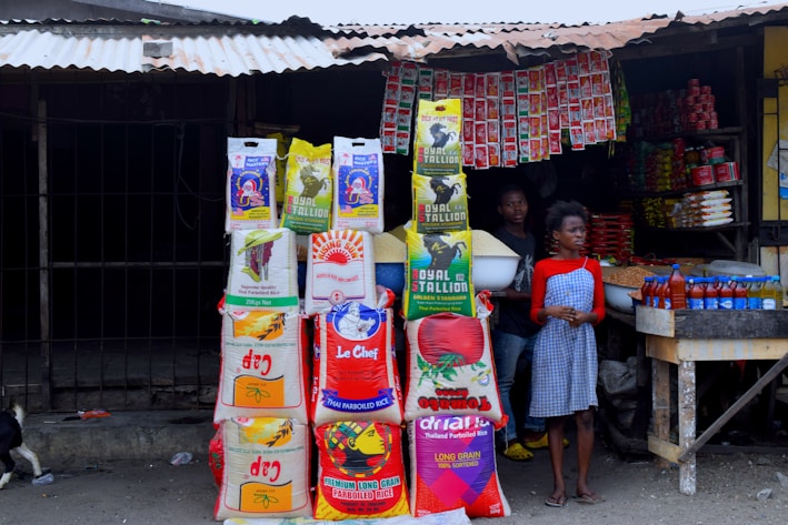 A small market stall displays various bags of rice, stacked in multiple columns. Two people, a young boy and a girl, are present; the boy holds a bowl of grains. Several red bottles are neatly arranged on a wooden table. Packs of hanging seasonings and instant noodles are visible in the background inside the dimly lit stall, with a metal roof overhead.