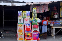 A small market stall displays various bags of rice, stacked in multiple columns. Two people, a young boy and a girl, are present; the boy holds a bowl of grains. Several red bottles are neatly arranged on a wooden table. Packs of hanging seasonings and instant noodles are visible in the background inside the dimly lit stall, with a metal roof overhead.
