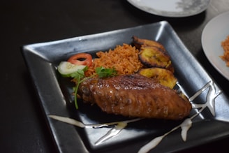 A black square plate contains a serving of jollof rice garnished with a slice of tomato and cucumber. Accompanying the rice are slices of fried plantain and a grilled chicken wing. The presentation is enhanced by a drizzle of sauce around the plate.