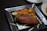 A black square plate contains a serving of jollof rice garnished with a slice of tomato and cucumber. Accompanying the rice are slices of fried plantain and a grilled chicken wing. The presentation is enhanced by a drizzle of sauce around the plate.