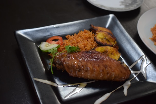 A black square plate contains a serving of jollof rice garnished with a slice of tomato and cucumber. Accompanying the rice are slices of fried plantain and a grilled chicken wing. The presentation is enhanced by a drizzle of sauce around the plate.