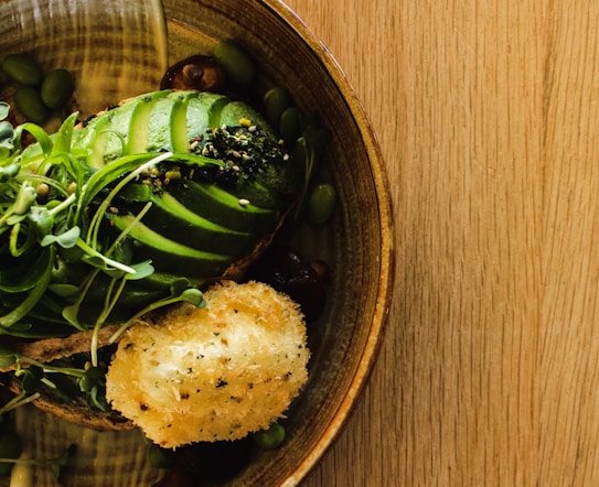 A wooden table holds a rustic ceramic bowl containing a beautifully arranged dish. Sliced avocado is layered neatly with microgreens sprinkled on top. Next to the avocado, a golden-brown breaded and fried piece appears to be a croquette. Edamame beans are scattered around the bowl, contributing to the composition.
