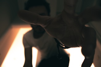A person reaching towards the camera, creating a sense of depth and movement. The background is softly illuminated, while the foreground is dominated by an outstretched hand. The lighting is low and dramatic, casting shadows that enhance the mysterious atmosphere.