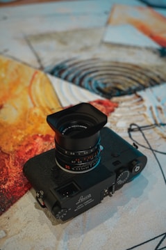 A vintage Leica camera with a prominent lens placed on a textured, abstract artwork. The artwork features swirls and splashes of warm colors, including yellow, orange, and red, which create an artistic backdrop for the camera.