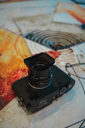 A vintage Leica camera with a prominent lens placed on a textured, abstract artwork. The artwork features swirls and splashes of warm colors, including yellow, orange, and red, which create an artistic backdrop for the camera.