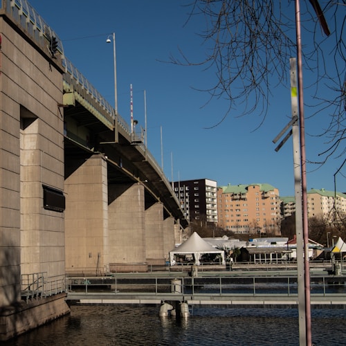 A large concrete bridge extends over a body of water with multiple support pillars. Below the bridge, there are several white tents set up near the water&rsquo;s edge. In the background, there are mid-rise residential buildings with varied colors. The sky is clear and blue, and there are leafless tree branches visible at the top right.
