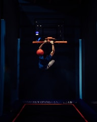 A person is hanging onto a basketball hoop with both hands while holding a bright orange ball. The setting appears to be a dark indoor gym or sports facility, with accent lighting that highlights the subject. The person is wearing a blue shirt and light-colored shorts.