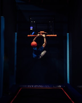 A person is hanging onto a basketball hoop with both hands while holding a bright orange ball. The setting appears to be a dark indoor gym or sports facility, with accent lighting that highlights the subject. The person is wearing a blue shirt and light-colored shorts.