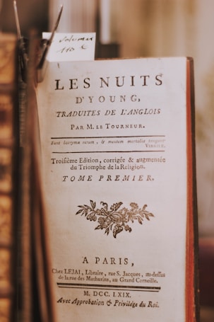 A close-up of an antique book page with text in French that includes the title 'Les Nuits d'Young.' The page appears to be aged with a decorative border and an ornate floral design near the bottom. The book appears to be positioned among other books with a visible price tag reading 'Volume 110€.'