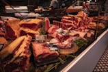 An assortment of meat cuts is displayed in a butcher shop's glass counter. The cuts vary in size and type, with visible marbling and fat. Price tags and labels are placed on some of the products, indicating their variety and cost. The counter is trimmed with green herbs for decoration. In the background, there are other shop elements like a cutting machine and a staff member preparing additional products.