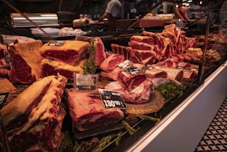 An assortment of meat cuts is displayed in a butcher shop's glass counter. The cuts vary in size and type, with visible marbling and fat. Price tags and labels are placed on some of the products, indicating their variety and cost. The counter is trimmed with green herbs for decoration. In the background, there are other shop elements like a cutting machine and a staff member preparing additional products.