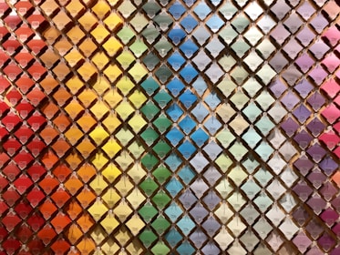 A wall filled with a neatly arranged array of colorful paint swatches. Each swatch is displayed in a diamond-shaped grid pattern, creating a rainbow gradient effect from warm reds and oranges on the left to cooler blues and purples on the right.
