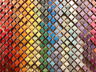 A wall filled with a neatly arranged array of colorful paint swatches. Each swatch is displayed in a diamond-shaped grid pattern, creating a rainbow gradient effect from warm reds and oranges on the left to cooler blues and purples on the right.