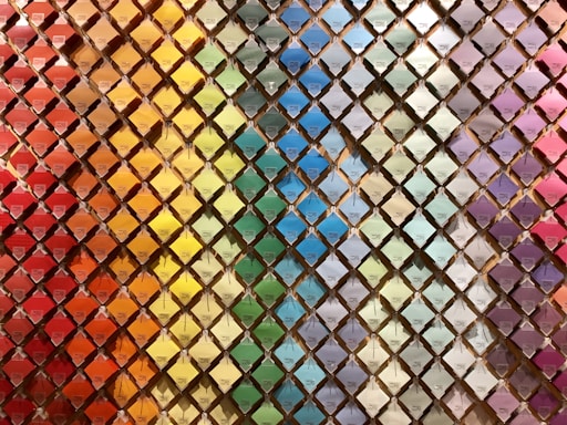 A wall filled with a neatly arranged array of colorful paint swatches. Each swatch is displayed in a diamond-shaped grid pattern, creating a rainbow gradient effect from warm reds and oranges on the left to cooler blues and purples on the right.
