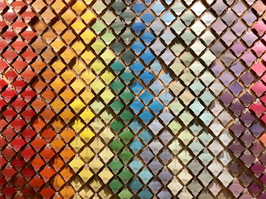 A wall filled with a neatly arranged array of colorful paint swatches. Each swatch is displayed in a diamond-shaped grid pattern, creating a rainbow gradient effect from warm reds and oranges on the left to cooler blues and purples on the right.