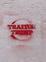 Red graffiti text reading 'TRAITOR TRUMP' is spray-painted on a rough, light-colored concrete wall.