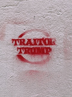 Red graffiti text reading 'TRAITOR TRUMP' is spray-painted on a rough, light-colored concrete wall.