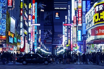 Vibrant cityscape featuring a busy urban street filled with people. Neon signs and advertisements cover the buildings, creating a lively atmosphere. A black car is parked in the foreground. Tall skyscrapers and illuminated storefronts are visible, with a mix of English and Japanese text on the signs.