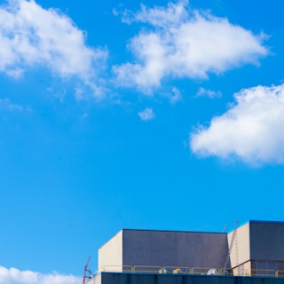 A modern building with a flat roof and a ladder extending to the top is set against a bright blue sky. Fluffy white clouds are scattered across the sky, adding contrast to the scene. The structure has clean lines and appears to be industrial or commercial in nature.