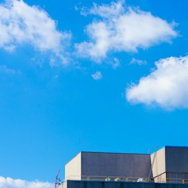 A modern building with a flat roof and a ladder extending to the top is set against a bright blue sky. Fluffy white clouds are scattered across the sky, adding contrast to the scene. The structure has clean lines and appears to be industrial or commercial in nature.