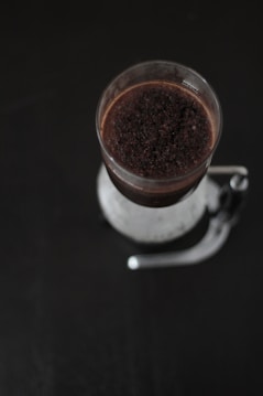 A top-down view of a French press coffee maker containing coffee grounds and water. The cylindrical glass vessel displays a dark brown mixture that contrasts against the sleek, black background, highlighting the brewing process.