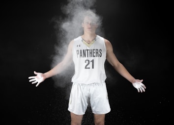 A basketball player in a 'Panthers' jersey marked with the number 21 stands with arms outstretched, surrounded by a cloud of chalk powder. The background is a stark black, emphasizing the white uniform and powder.