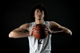 A basketball player with curly hair is wearing a white jersey with the number 12. He is holding a basketball with both hands close to his chest. The background is dark, highlighting his focused expression and muscular build.