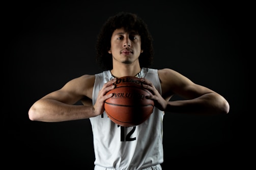 A basketball player with curly hair is wearing a white jersey with the number 12. He is holding a basketball with both hands close to his chest. The background is dark, highlighting his focused expression and muscular build.