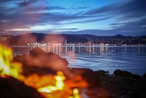 A vibrant cityscape at dusk across a calm body of water, with city lights reflecting on the surface. In the foreground, blurred flames add warmth and contrast against the cool tones of the water and sky. Hills silhouette the city skyline, and scattered clouds add texture to the blue evening sky.