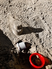 A small sandcastle mold sits on a textured surface of sand. Beside it is a red plastic ring and a small white plastic cup partially embedded in the sand. Sunlight creates shadows, adding depth to the image.