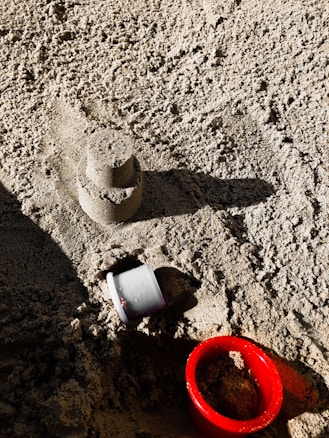 A small sandcastle mold sits on a textured surface of sand. Beside it is a red plastic ring and a small white plastic cup partially embedded in the sand. Sunlight creates shadows, adding depth to the image.