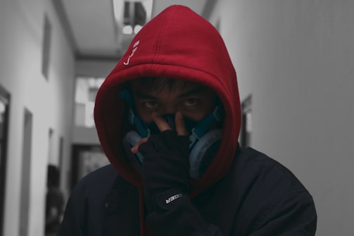 A person wearing a red hooded sweatshirt is standing in a narrow hallway. The individual has dark hair and is wearing a blue respirator mask over their mouth and nose. Their eyes are partially visible and focused forward, giving a mysterious vibe. The background is blurry, and the lighting is dim, hinting at a moody or tense atmosphere.