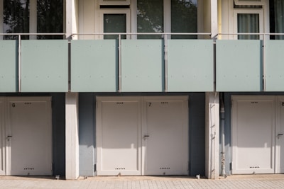 A series of closed garage doors numbered 6, 7, and 8 are positioned side by side beneath a modern apartment building. Above the doors, there is a glass balcony with a light green frosted panel railing and windows with shades partially drawn. The setting is likely urban and has a clean, minimalistic architectural design.