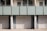 A series of closed garage doors numbered 6, 7, and 8 are positioned side by side beneath a modern apartment building. Above the doors, there is a glass balcony with a light green frosted panel railing and windows with shades partially drawn. The setting is likely urban and has a clean, minimalistic architectural design.